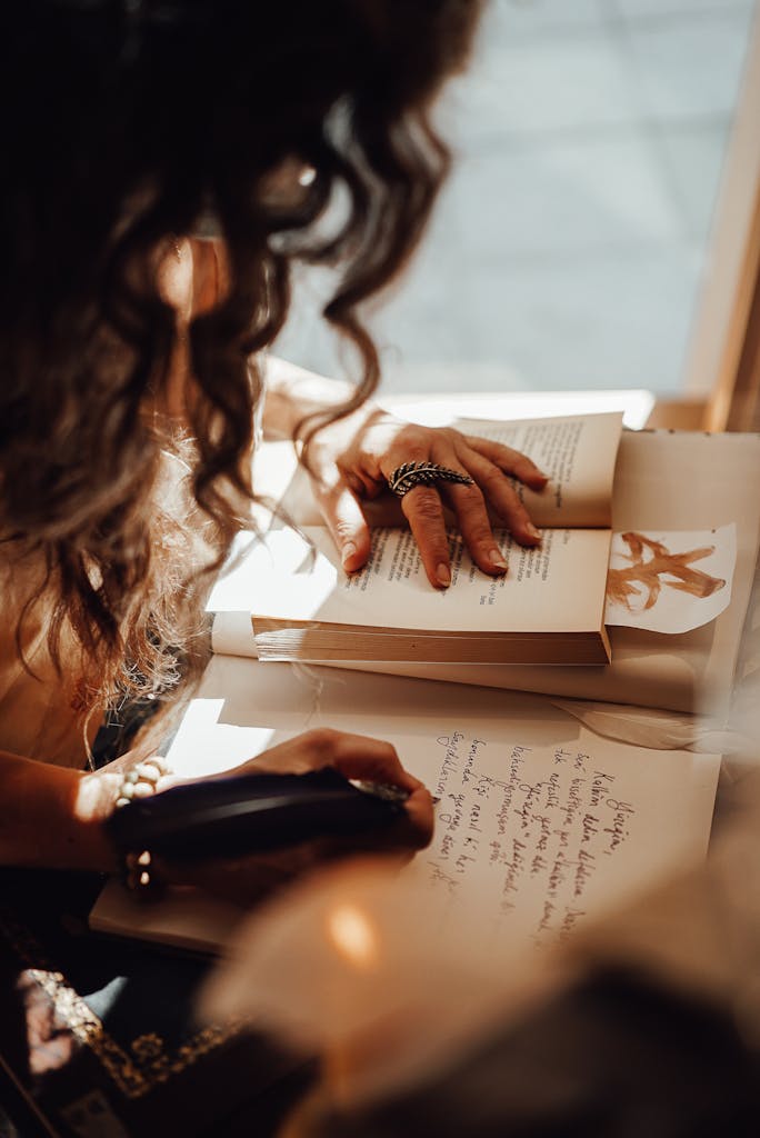 Crop anonymous female with dark hair sitting near window at retro writing cabinet while taking notes with feather in vintage store